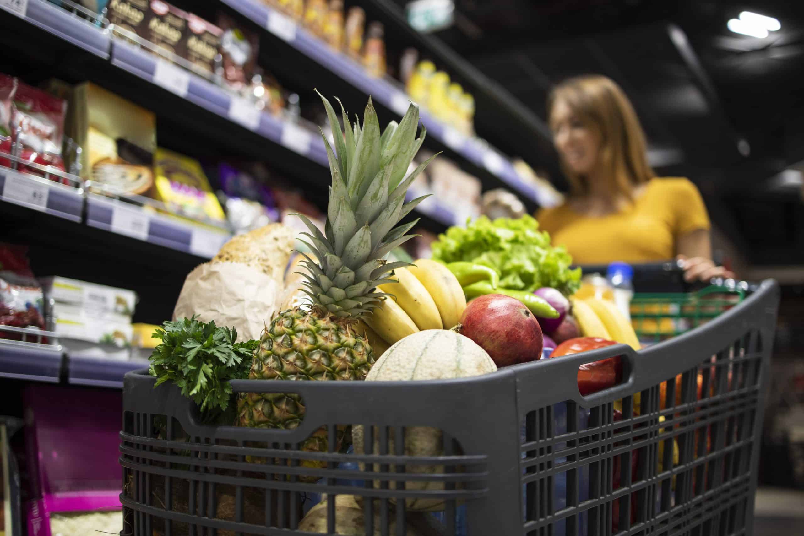 Close up view of shopping cart overloaded with food while in background female person choosing products.